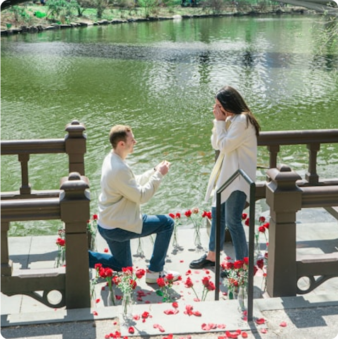 Photo d'un homme à genou devant une femme entourée de rose et qui la demande en mariage au bord d'un lac lors de fiançailles