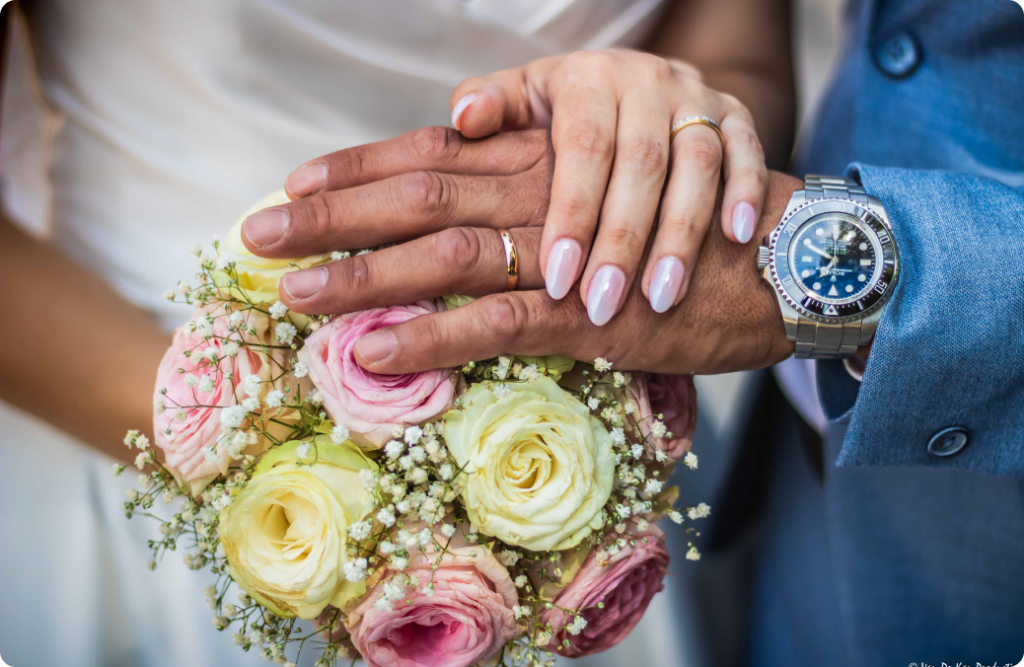 Photo des mains de mariés homme et femme posées l'une sur l'autre sur un bouquet de fleurs avec leur deux alliances